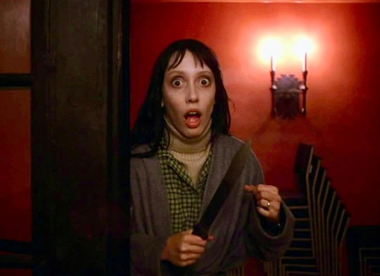 A woman with wide eyes and an open mouth, appearing shocked or scared, stands in a dimly lit room holding a knife. She wears a green checkered shirt, a grey cardigan, and has dark hair. The background features red walls, a staircase, and a lit sconce.