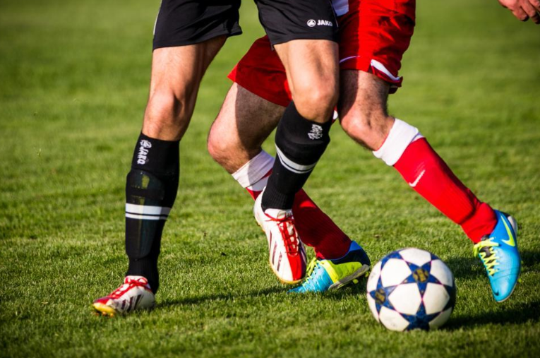 Two soccer players wearing black and red shorts, respectively, tussling for control of a soccer ball on a grassy field. Both players are in motion, with one wearing black socks and the other red socks and blue shoes. The ball has a star pattern.