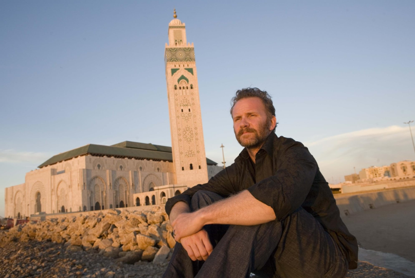 A man with a beard and wearing a dark shirt sits on a rocky ground, with a large mosque featuring a tall minaret in the background. The sky is clear and the sun casts a warm light over the scene.