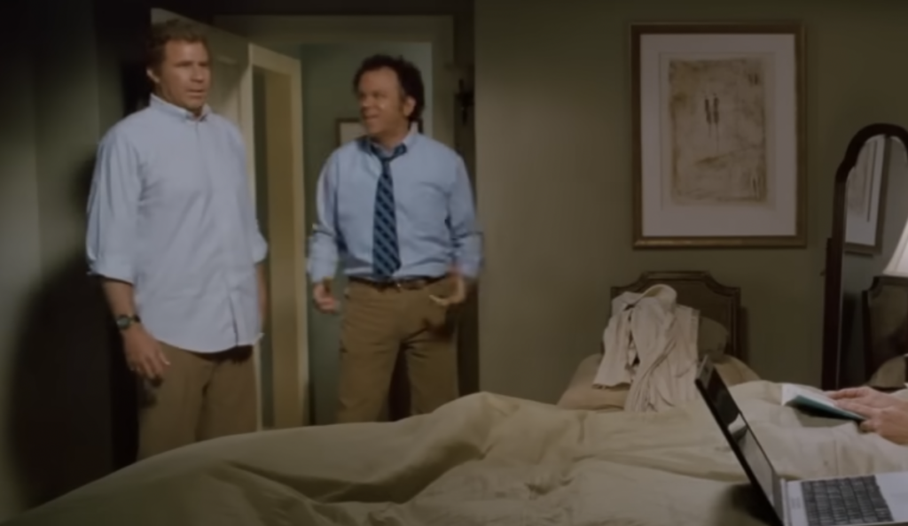 Two men in button-down shirts and khakis, one with a tie, stand in a bedroom. One looks surprised, while the other is talking. An unmade bed is in the foreground with a laptop and a person sitting with a book. A framed picture hangs on the wall.