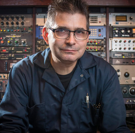 A person wearing glasses and a navy blue coverall is sitting with various audio equipment and controls in the background. The person has a neutral expression and has short, dark hair. A pen and a small notebook are visible in the front pocket of the coverall.