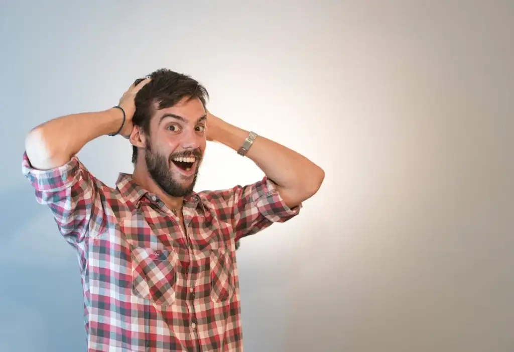 A man with a beard and a plaid shirt stands against a plain background with a surprised and excited expression. He has both hands on his head and his mouth open wide, as if reacting to something incredible.