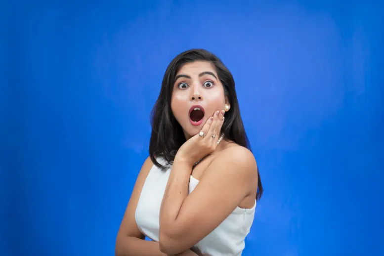 A woman with long dark hair and wearing a white sleeveless top is standing against a blue background. She has an expression of surprise with wide eyes and an open mouth, her left hand touching her cheek.