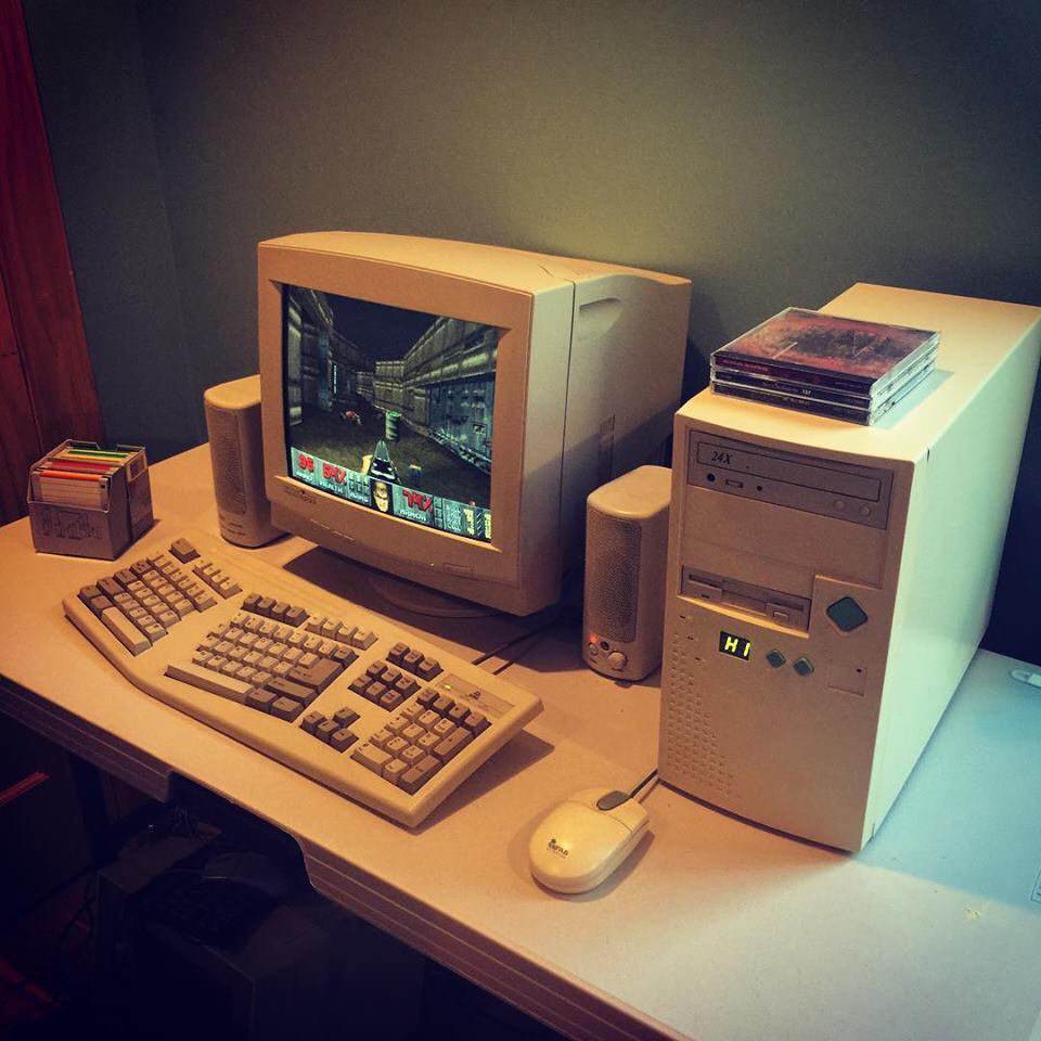 A vintage desktop computer setup with a CRT monitor displaying a retro first-person shooter game. The setup includes a beige keyboard, mouse, speakers, floppy disks in a box, and a stack of CDs on the PC tower. The computer is placed on a white desk.