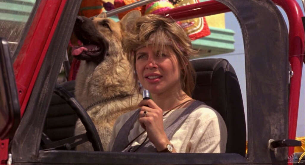 A woman with short, tousled blonde hair holds a walkie-talkie while sitting in the driver's seat of an open-top vehicle. A German Shepherd dog is sitting beside her, panting with its tongue out. The background shows bright, colorful details.