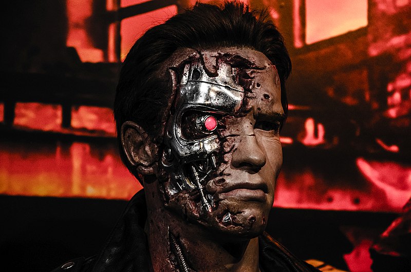 A close-up of a figure resembling a battle-damaged robot with a human-like face, revealing a metallic skull and a glowing red eye on one side. The background features an industrial setting with a reddish glow.