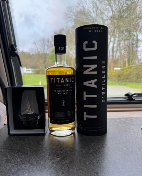 A bottle of Titanic Distillers Irish whiskey stands next to its black cylindrical packaging. A small box, containing a glass etched with a ship, is also placed beside the bottle. The background shows a blurred outdoor scene through a window.