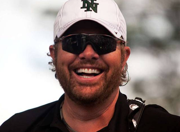 A smiling man with a beard wears dark sunglasses and a white baseball cap with a green logo. He has a dark shirt on and carries a black strap over his shoulder. The background is blurry, highlighting his cheerful expression.