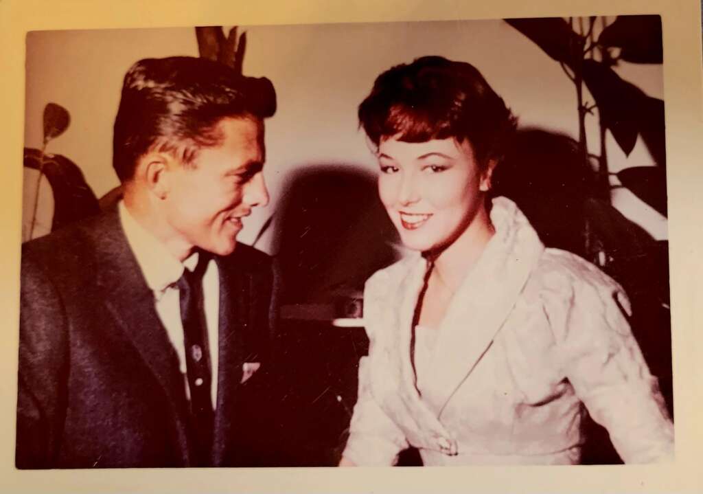 A sepia-toned photograph shows a man and a woman. The man, dressed in a suit and tie, is smiling and facing the woman. The woman, wearing a light-colored dress, is smiling at the camera. They are indoors, and there are plants in the background.