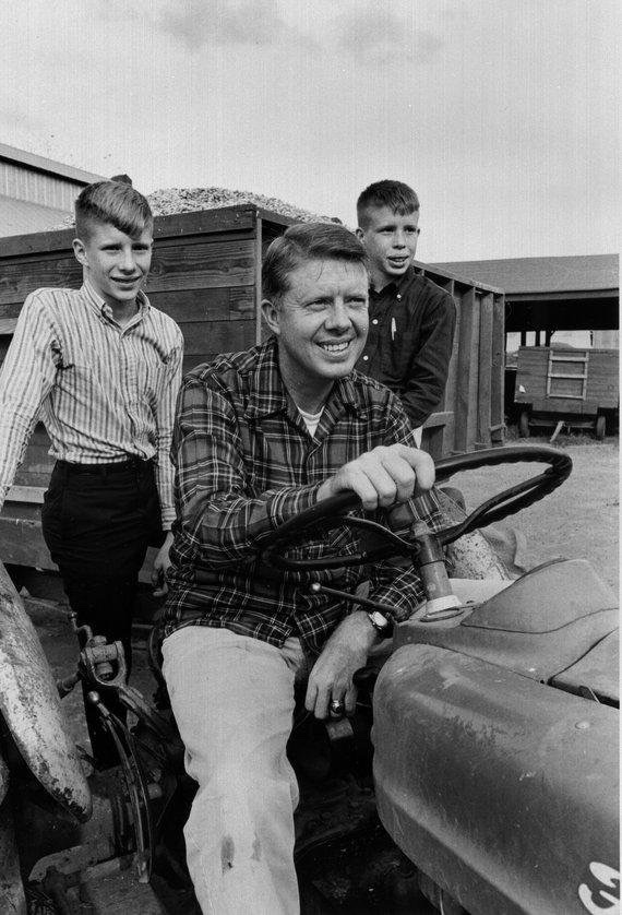 A black and white photo shows a smiling man in a flannel shirt sitting on a tractor, holding the steering wheel. Two boys stand behind him, one on each side, both dressed in casual clothes. They are outdoors with a wooden structure and trailer visible in the background.