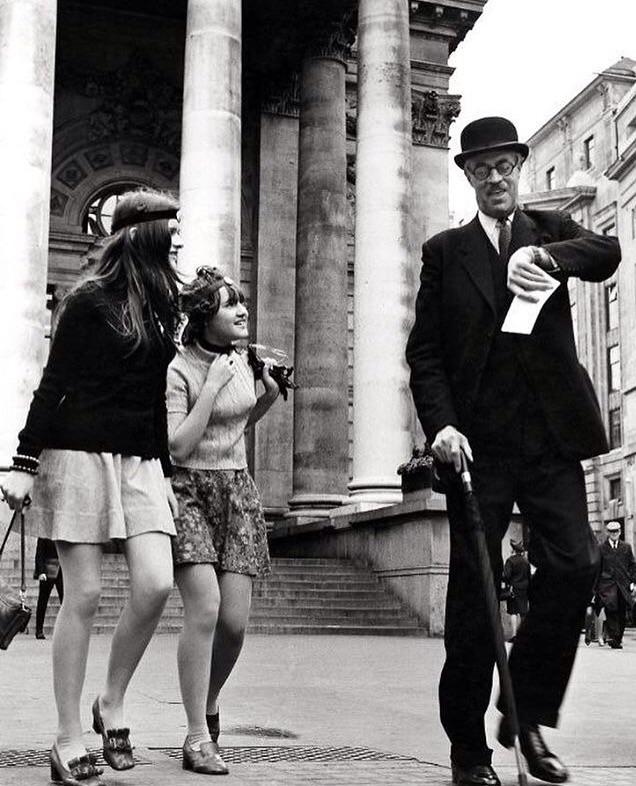 A black and white photograph showing an elderly man wearing a bowler hat and using a cane, appearing to dance on a city street. Two young women, dressed in 1960s fashion, are smiling and enjoying his performance in front of a grand building with large columns.