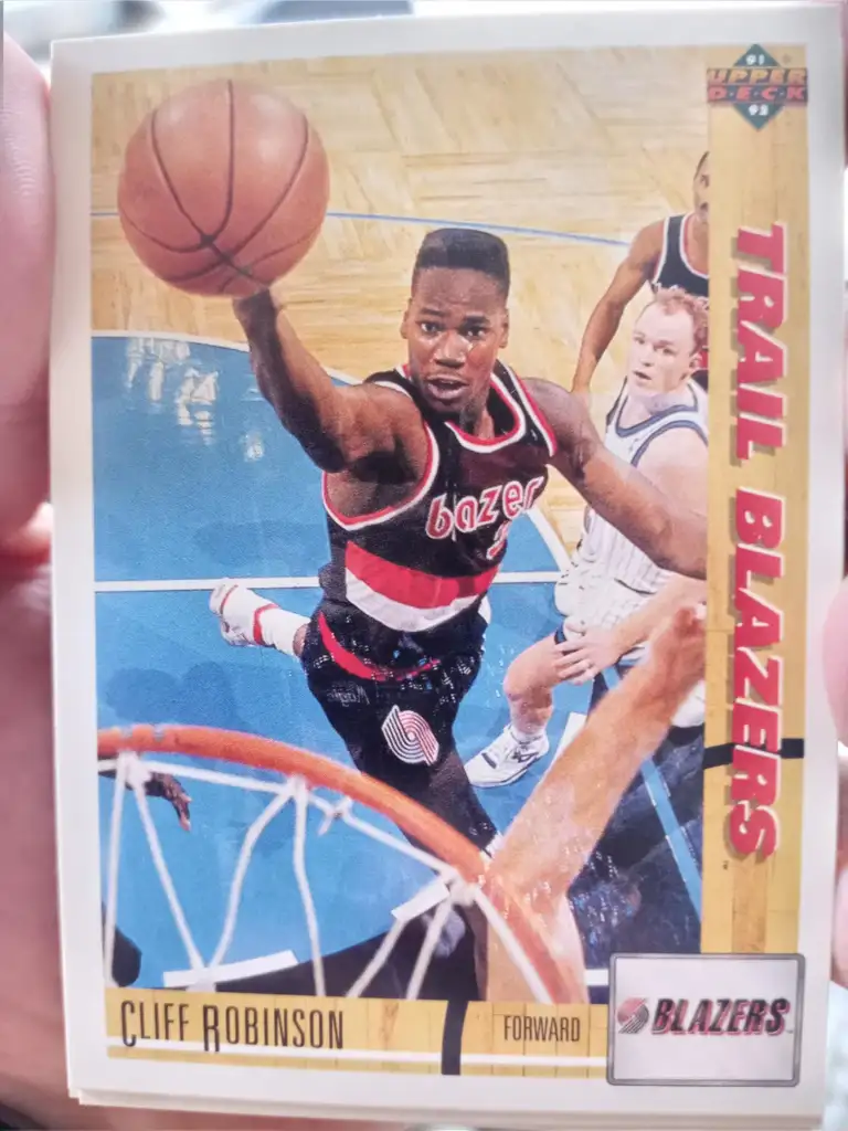 A trading card featuring basketball player Cliff Robinson of the Portland Trail Blazers. He is shown in action, wearing a red and black Blazers uniform and jumping towards the basket with the ball. The card has a yellow border, "Trail Blazers" text, and team logos.