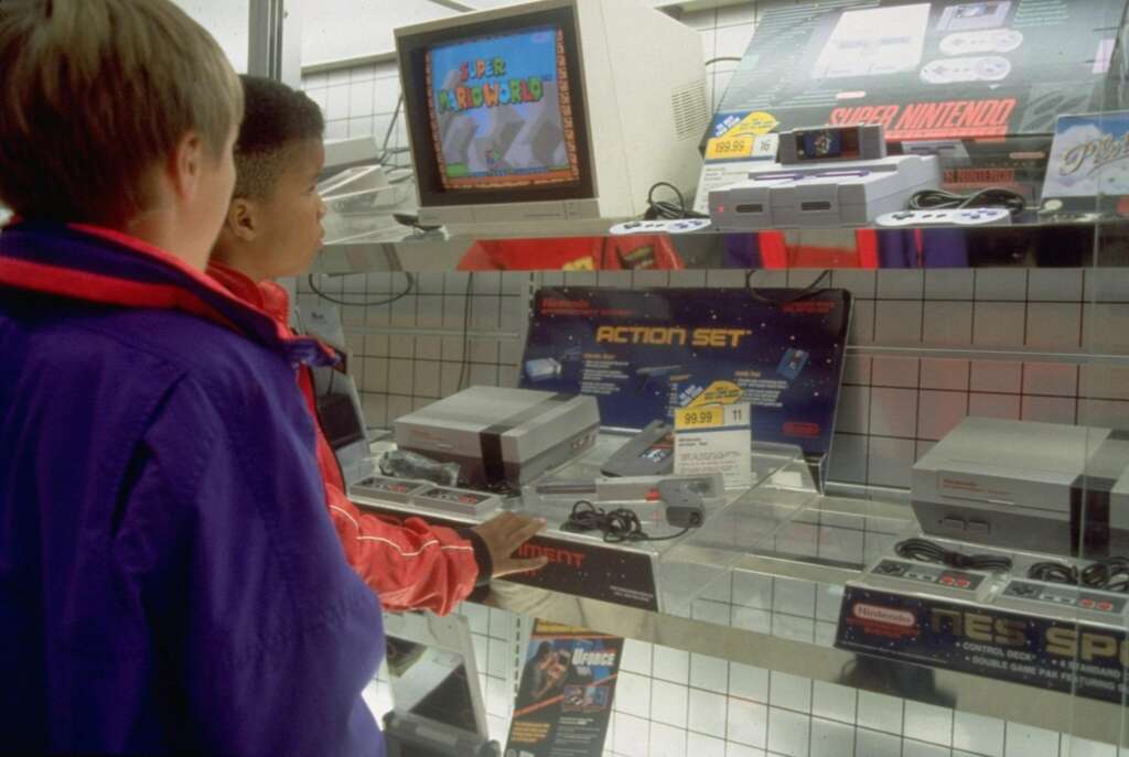 Two children are in a store, looking at a video game display. The display features NES and Super NES consoles, controllers, and games. An older style television shows "Super Mario World" being played. The children appear to be captivated by the display.