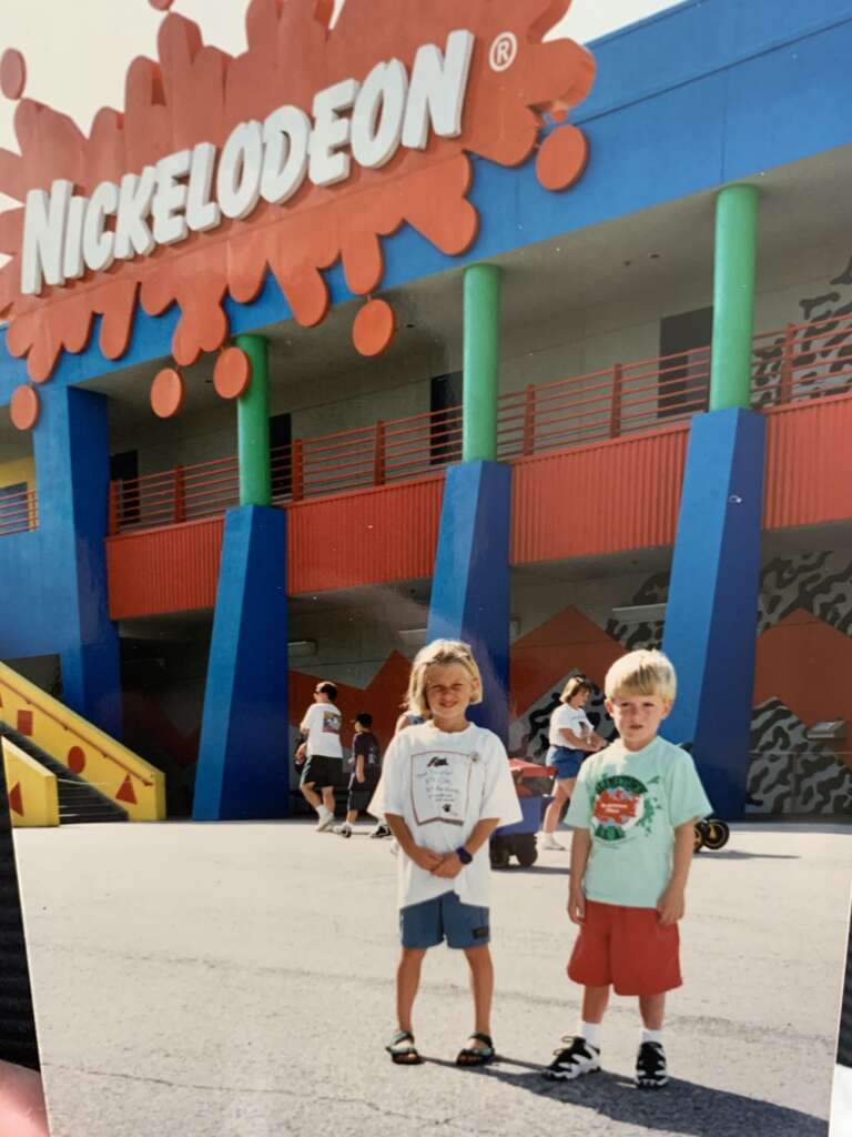 Two children stand in front of a colorful Nickelodeon building, smiling at the camera. The building features vivid blue pillars, green accents, and a large orange Nickelodeon sign with splat design. People walk in the background under a sunny sky.