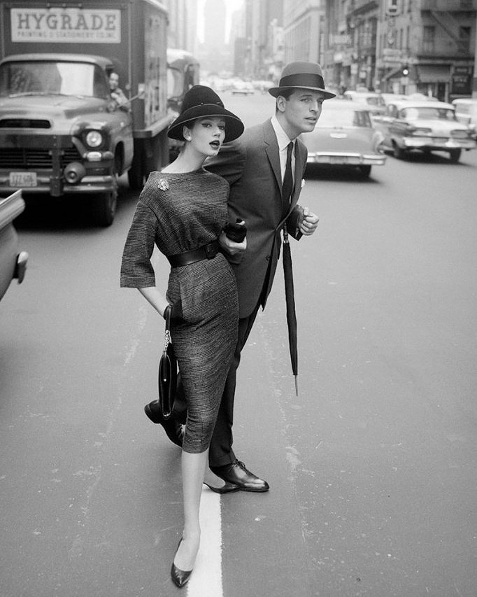 A black and white photograph depicts a stylishly dressed woman and man crossing a busy urban street. The woman wears a fitted dress and hat, accessorized with gloves and a bag. The man, wearing a suit and hat, holds an umbrella. Vintage cars and buildings are visible.