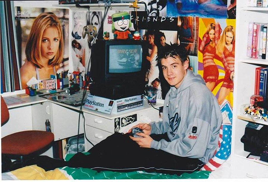 A teenager sits on a bed playing PlayStation in a room decorated with various posters and figurines. The room has a cluttered desk, a small TV displaying a game, and vibrant posters of TV shows and celebrities on the walls.