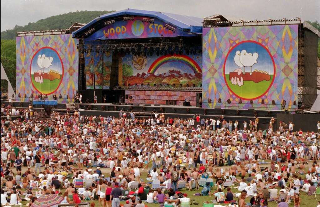 A large outdoor festival stage with "Woodstock" written on a sign and colorful decorations. Two large banners feature peace symbols with a white dove on a guitar. A crowd of people is gathered in front, some sitting on the grass, others standing, enjoying the event.