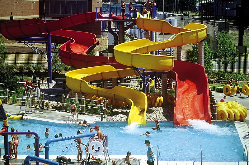 A vibrant water park featuring three winding water slides—red, orange, and yellow—plunging into a swimming pool. People are enjoying the slides and swimming in the pool. Tubes are stacked nearby, and the area is surrounded by greenery and buildings.