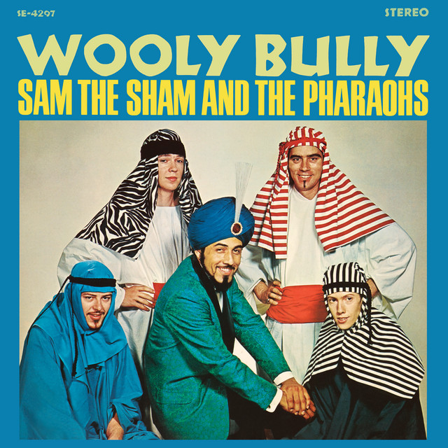 Album cover for "Wooly Bully" by Sam the Sham and the Pharaohs. A group of five men, dressed in a mix of traditional Middle Eastern attires and Western outfits, smile and pose against a blue background. The top text displays the album and artist name in bold yellow letters.