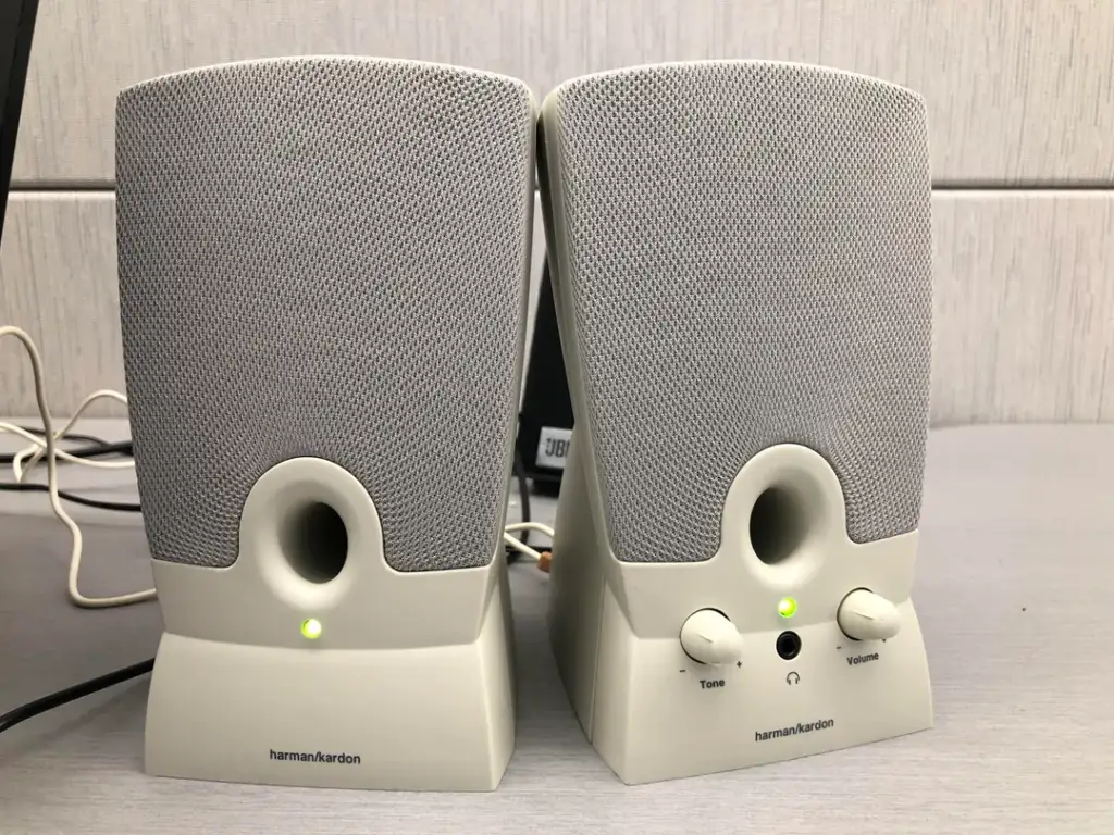 A pair of Harman Kardon desktop speakers sit side by side on a grey surface. The right speaker has a green indicator light and two knobs labeled "Tone" and "Volume." The left speaker has a matching design. Background includes grey partition and connecting cables.