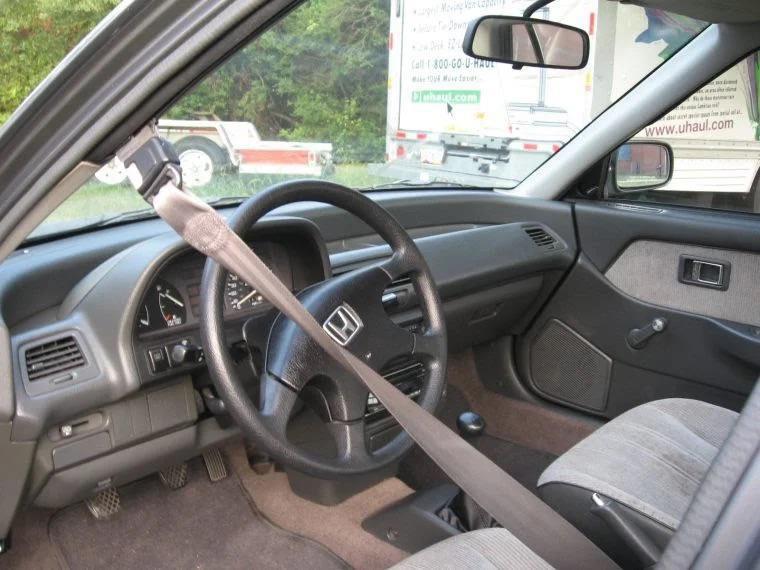 Interior of a Honda car showing the driver's side with a steering wheel, dashboard, speedometer, and climate controls. The passenger seatbelt is fastened. The view includes the interior door panel and side mirror with a U-Haul trailer visible through the window.