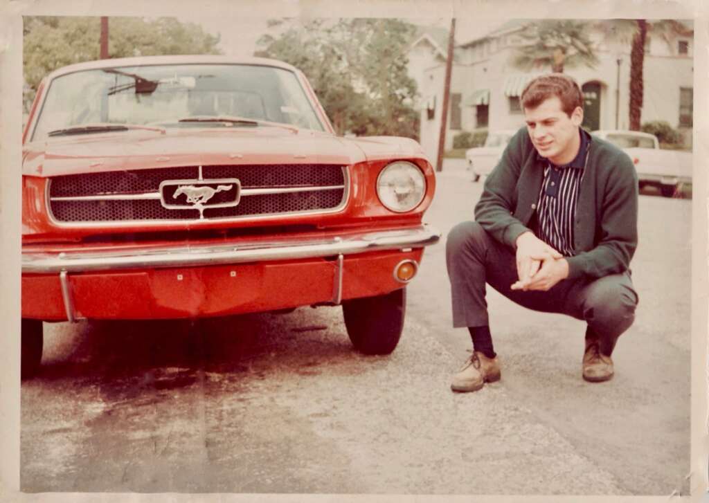 A man squats next to a red vintage Ford Mustang car on a residential street. He is wearing a green cardigan over a striped shirt, dark pants, and brown shoes. Trees and houses are visible in the background. His expression is contemplative.