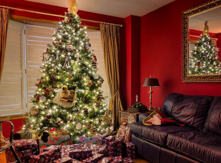 A decorated Christmas tree adorned with lights, ornaments, and an angel on top stands in a warmly lit living room with rich red walls. Presents wrapped in festive paper are placed underneath the tree. A dark leather couch and a gold-framed mirror are nearby.