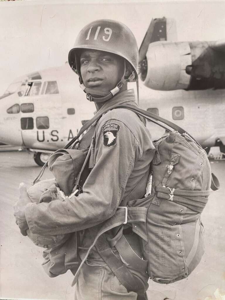 A soldier stands in front of a U.S. Air Force aircraft, wearing a helmet with the number 119 and a parachute on his back. He is dressed in military fatigues and looks into the camera, his right hand holding a strap of his gear. A "101st Airborne" patch is visible on his sleeve.