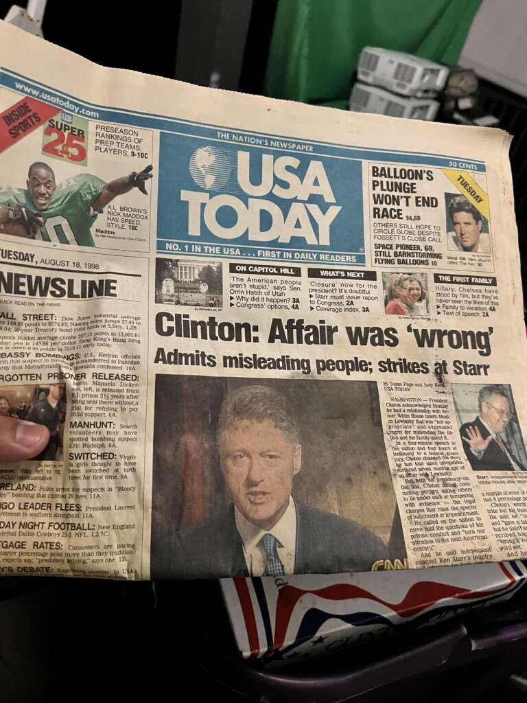 A person holds a USA Today newspaper with the headline: "Clinton: Affair was 'wrong' - Admits misleading people; strikes at Starr." The date on the newspaper is Tuesday, August 18, 1998. The front page also features various other news articles and a photo of Bill Clinton.