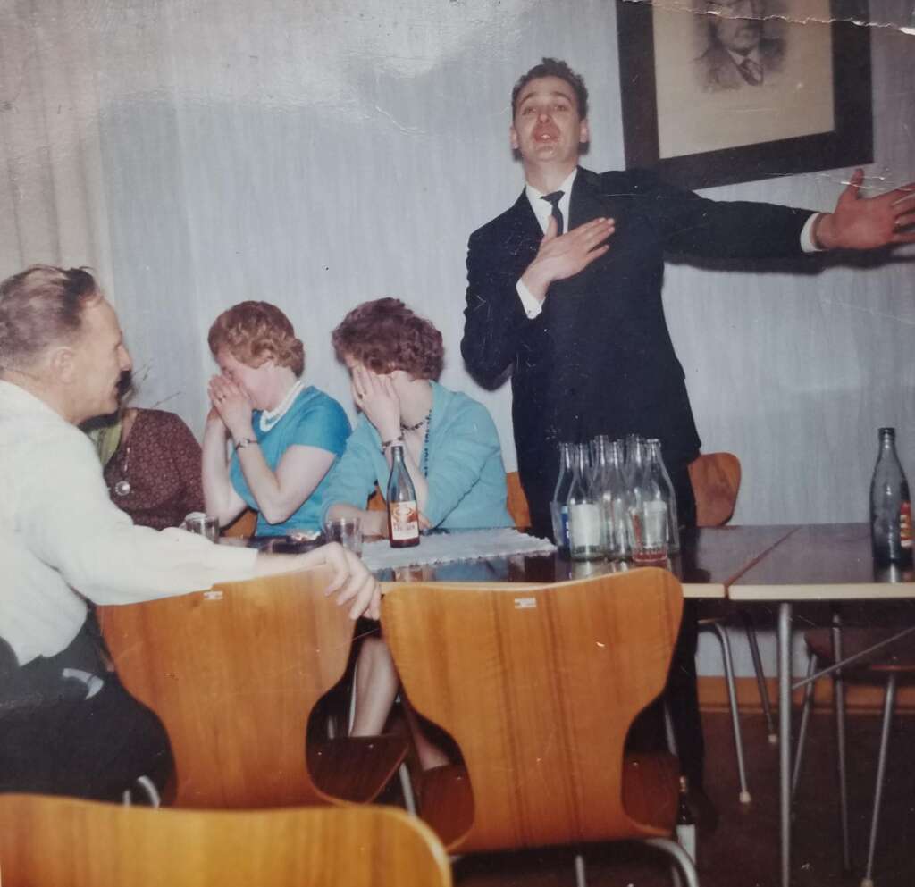 A man in a suit stands with one hand on his chest and the other arm extended, speaking or singing passionately in a room with several seated people. Two women cover their faces while another man and woman look on. Bottles and glasses are on the tables in front of them.