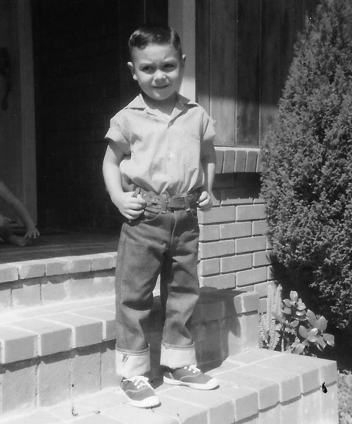 A young boy stands on brick steps in front of a house. He's wearing a short-sleeve shirt, cuffed jeans, and sneakers. His hair is neatly combed, and he has a slight smile on his face. The background includes part of a wall, a doorway, and some foliage.