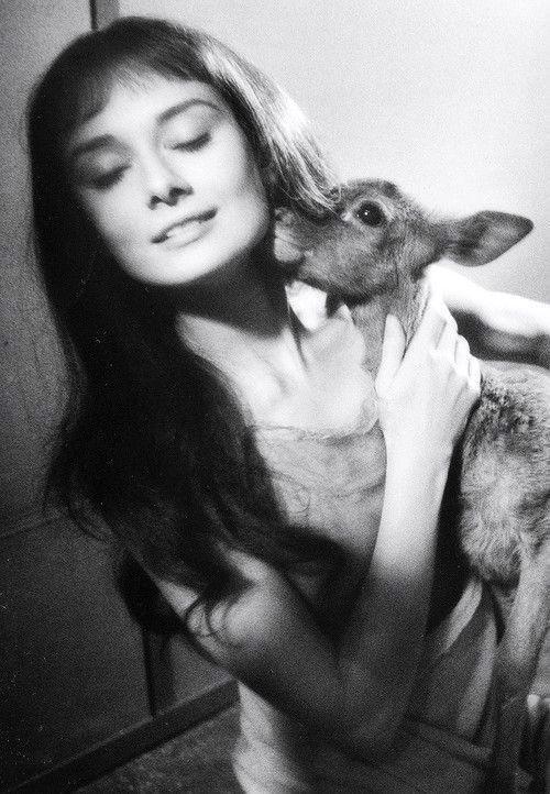 A woman with long hair smiling gently while holding a fawn close to her face. The fawn is nuzzling against her, and the scene is illuminated softly, creating a warm and intimate atmosphere. The image is in black and white.