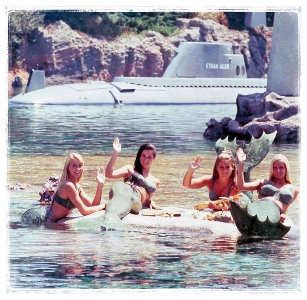 Four women dressed as mermaids sit on rocks in a body of water, smiling and waving. They have mermaid tails and are wearing tops that match the tails. In the background, a submarine with the name "Ethan Allen" can be seen near a rocky shore.