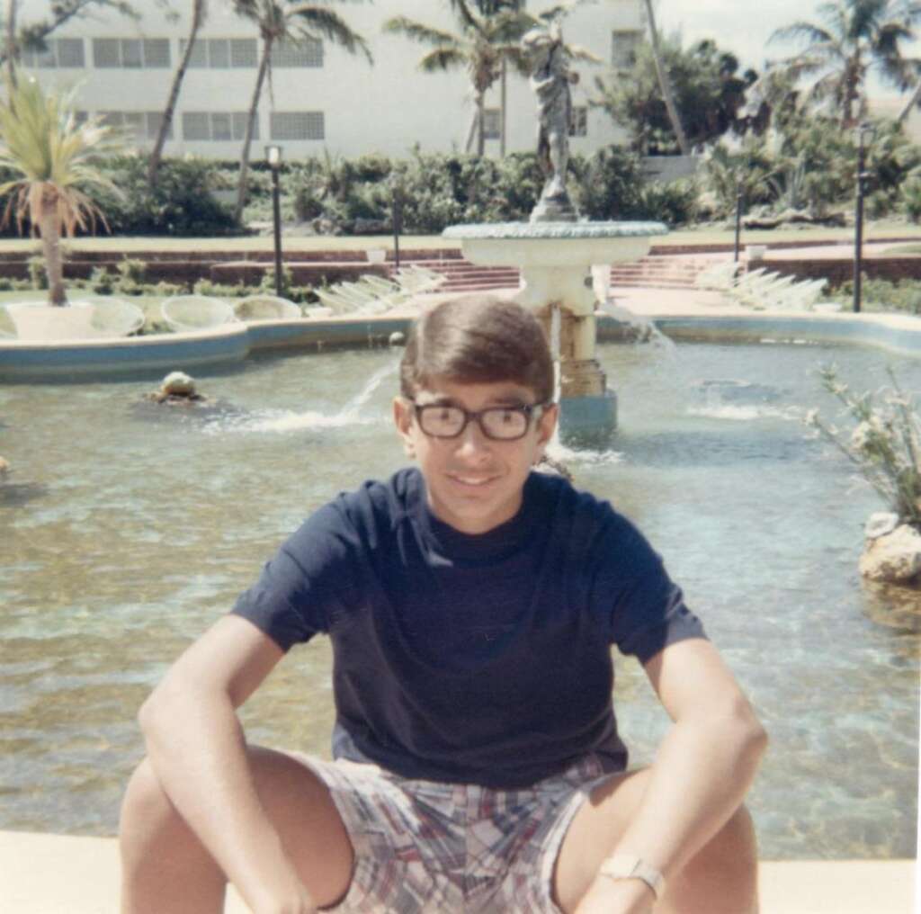 A young person with glasses, wearing a navy blue t-shirt and checkered shorts, sits by a decorative pond with a fountain. In the background, there are palm trees and a building. The day appears sunny and bright.