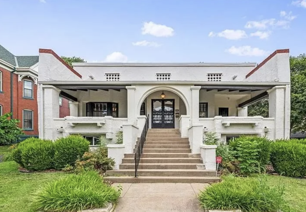 A large white bungalow-style house with a prominent arched entryway and an expansive front veranda. Wide steps with a central black handrail lead up to the front door. The house is surrounded by well-manicured bushes and grass, with a neighboring red brick building visible.