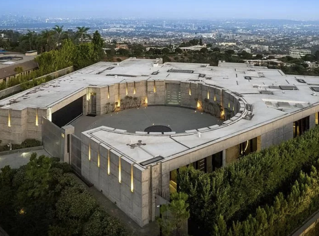 An aerial view of a large, modern concrete mansion with a circular courtyard in the center. The mansion is surrounded by lush greenery and offers a clear view of a sprawling cityscape in the background, under a hazy blue sky.