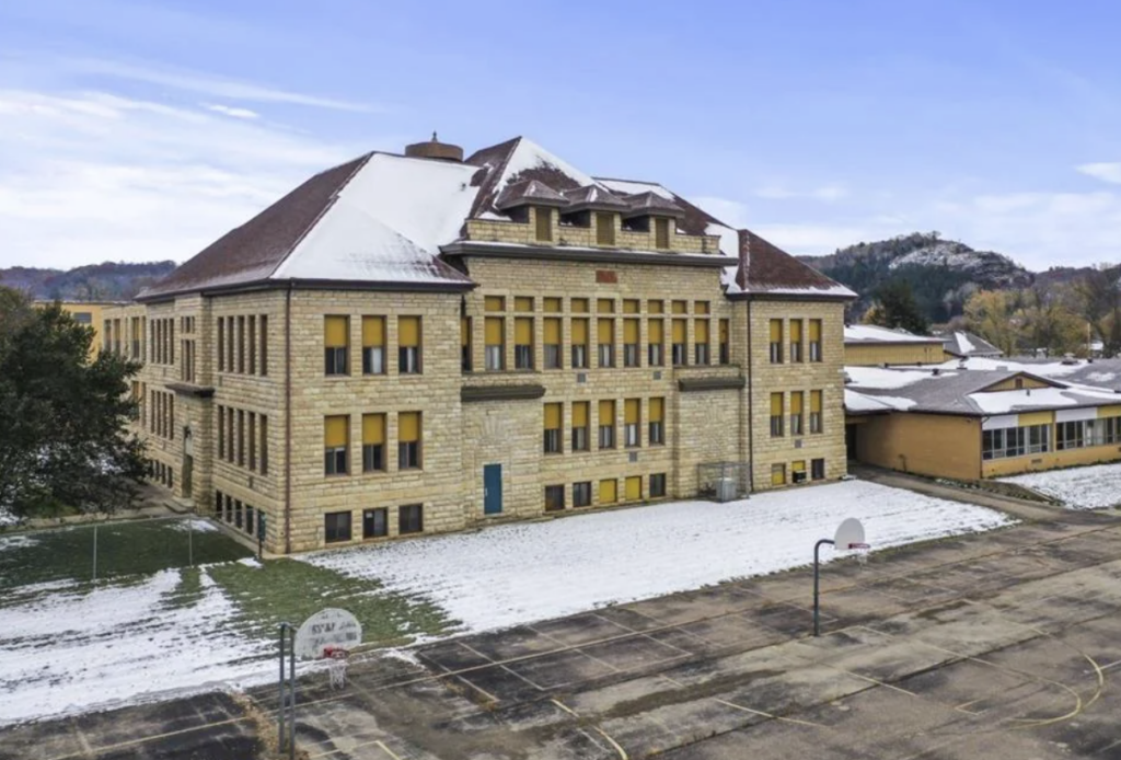 A large, old-fashioned stone building with snow on its roof and patches of snow on the surrounding grass. The building has multiple stories with numerous yellow windows. In the foreground, there is a large paved area with basketball hoops. Hills are visible in the background.