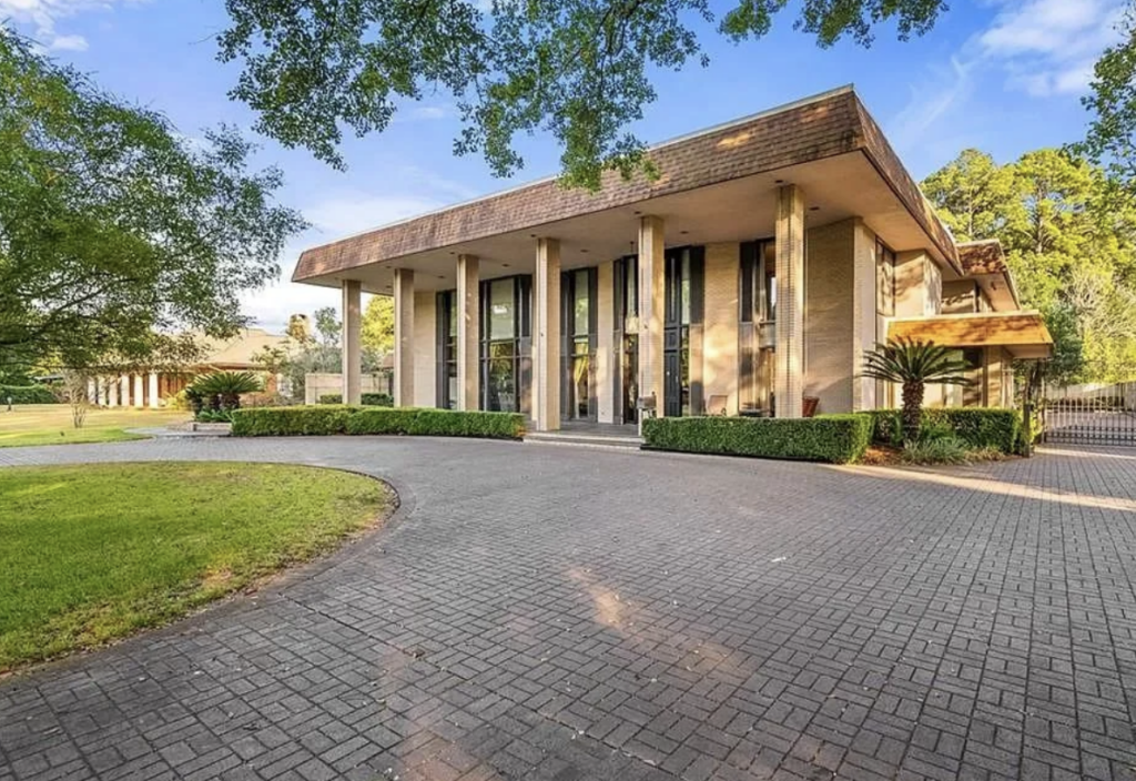 A modern, single-story building with a broad flat roof and multiple vertical pillars at the entrance. The structure has large glass windows, and is surrounded by trees, manicured shrubs, and a paved driveway that leads to the front door. A blue sky is visible above.