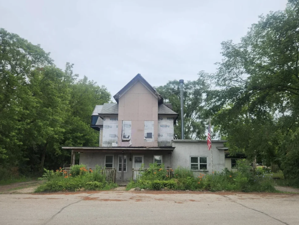 A two-story, partially renovated house with a slanted roof stands among green trees. The façade is partially covered with house wrap. An American flag is displayed on the right side, and there are garden plants and flowers in front of the building. The sky is overcast.