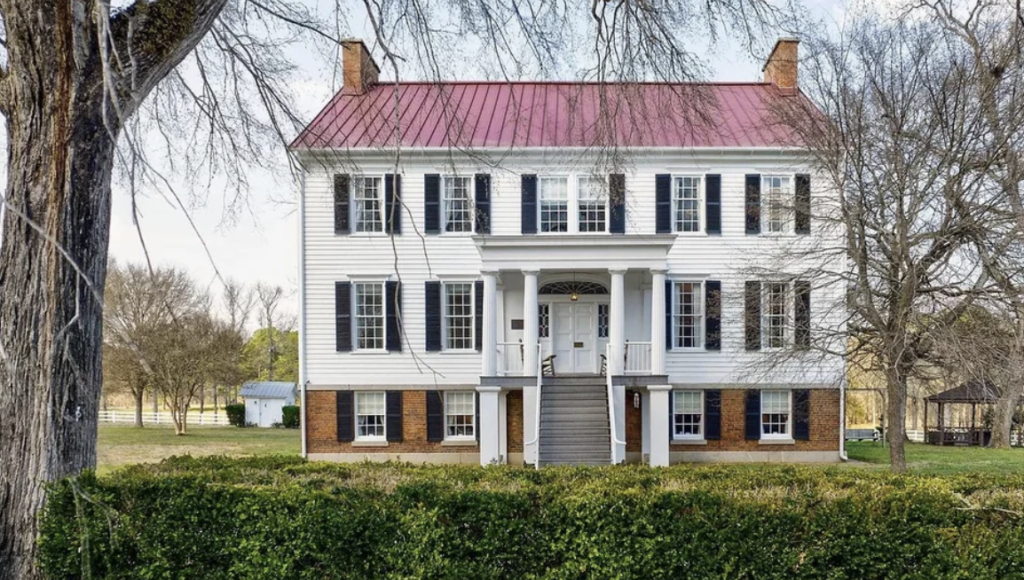 A large, two-story colonial-style house with a red metal roof, white siding, and black shutters. It has a central porch with columns and a staircase leading to the entrance. The yard is surrounded by bare trees and a trimmed green hedge in the front.