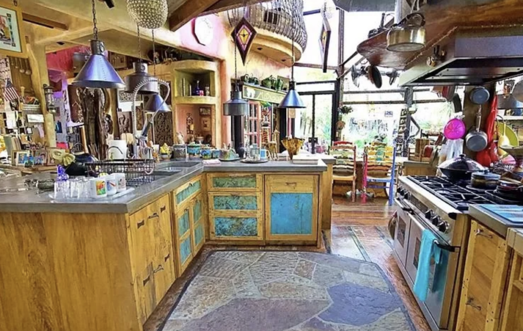 A colorful, eclectic kitchen features rustic wooden cabinets with blue panels, a large stone-patterned floor mat, and modern hanging lights. Shelves are filled with various knick-knacks, and large windows at the back let in natural light, showcasing a lively room.