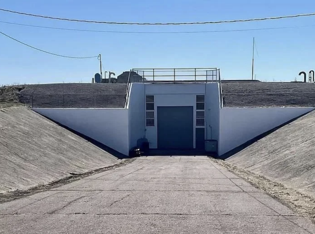 A concrete bunker entrance is built into a mound of earth with a wide, paved pathway leading to it. The entrance has a large, gray metal door and is flanked by small windows. Electrical wires and equipment are visible on top of the mound under a clear blue sky.