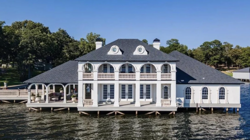 A large, two-story house built over a lake with white walls and prominent arches on the upper level. It has a covered gazebo area on the left, surrounded by trees, and an expansive water view in the foreground. The blue sky is clear and sunny.