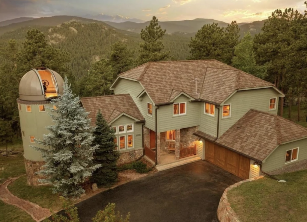 A large, greenish-gray house with a brown roof is set among dense trees and mountains at sunset. The house features multiple stories, a round tower with a conical roof on the left, and an expansive driveway with a two-car garage on the right.