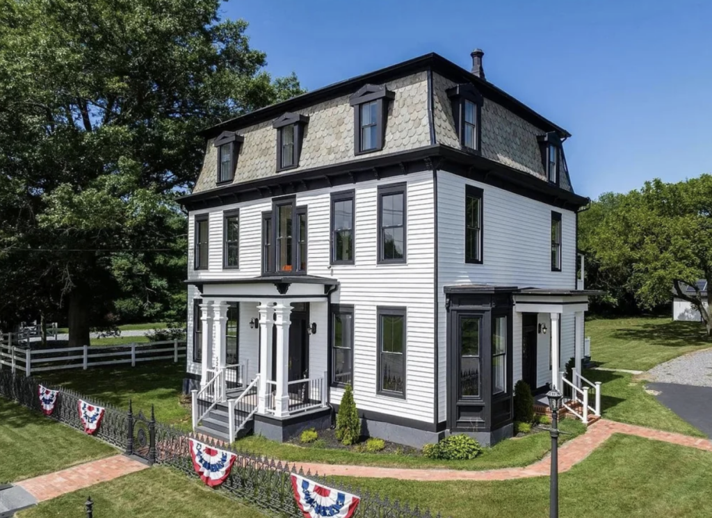 A white, two-story house with black trim and roofing, featuring a mansard roof and decorative dormer windows. The porch has classic columns, and a well-maintained lawn surrounds the home. American flag buntings hang on the fence in front, and trees are visible in the background.