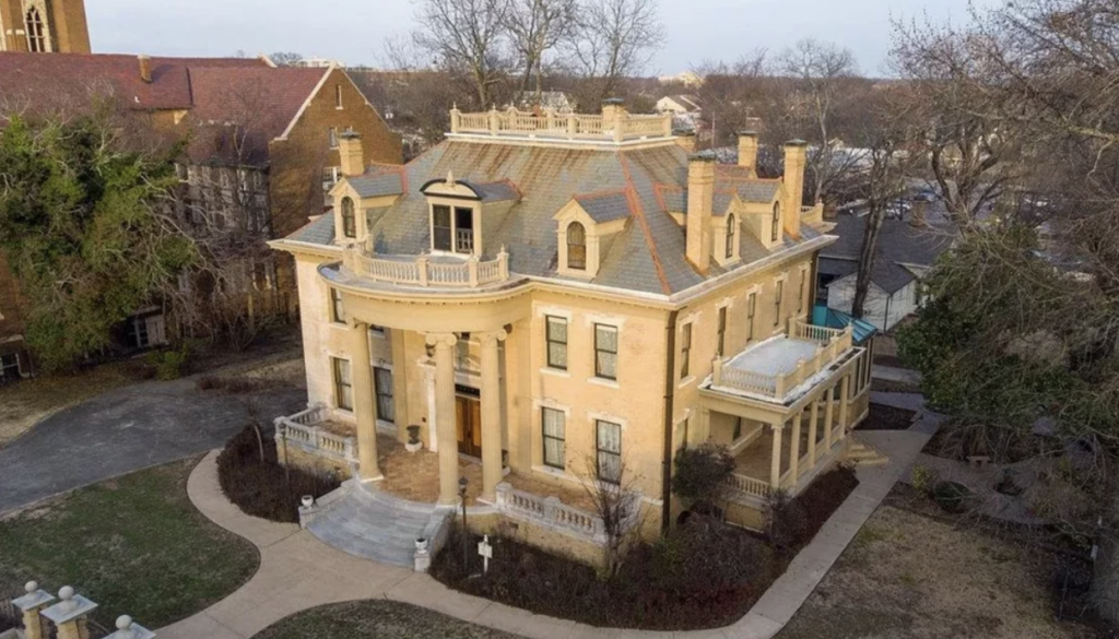 An aerial view of a large, historic mansion with beige walls and red chimney tops. The house has a grand, columned entrance and a semicircular terrace. Trees and neighboring buildings surround it, and a well-kept lawn is in front.