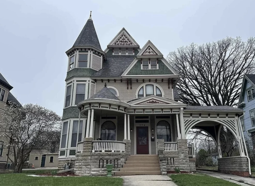 A grand Victorian house with a turret, multiple gables, and intricate woodwork. It features a wraparound porch, arched entrances, and a mix of stone and wood exterior. The house is set against a cloudy sky with bare trees and adjacent homes visible.