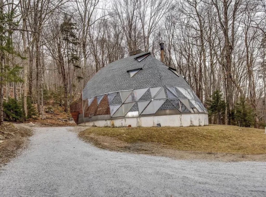 A unique dome-shaped house with triangular windows and a shingled roof stands in a wooded area. The house has a mix of shingled and brick exterior. A gravel driveway leads to the entrance, surrounded by bare trees and patches of grass.