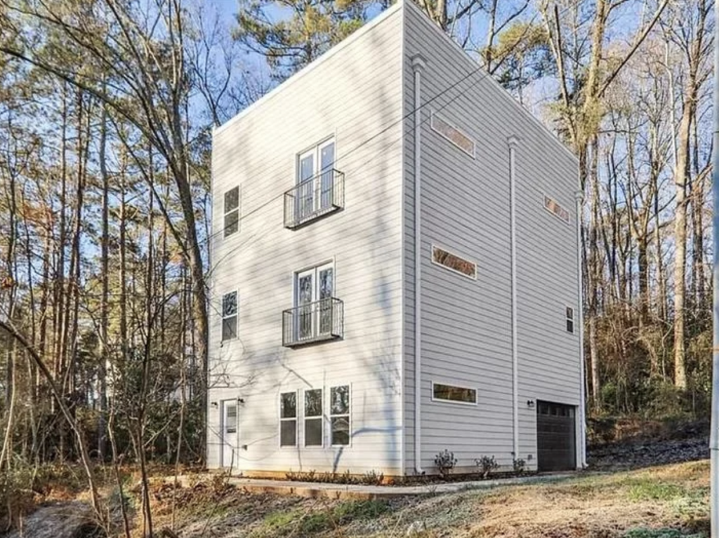 A tall, narrow, modern white house with a minimalist design stands in a wooded area. It has multiple small rectangular windows and two small balconies on the upper floors. The house features a garage on the lower level and a minimalist front entrance. Trees surround the property.