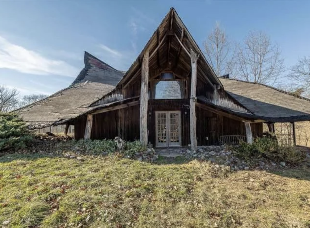 A unique, rustic wooden house with a tall, pointed roof and wide eaves. The structure features large wooden beams supporting the entrance. The surrounding grass and sparse trees suggest a rural setting, with clear blue skies above.